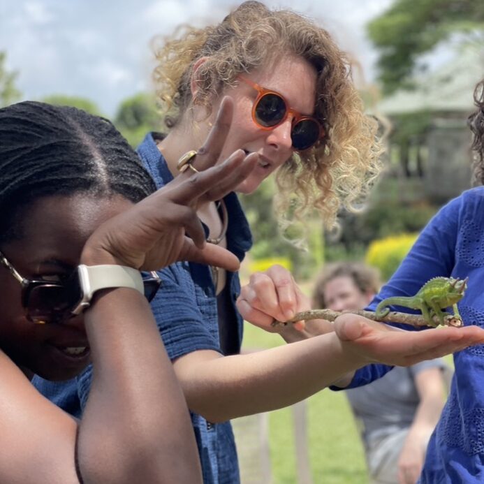A woman holds a lizard in her palm with another woman in the foreground gesturing